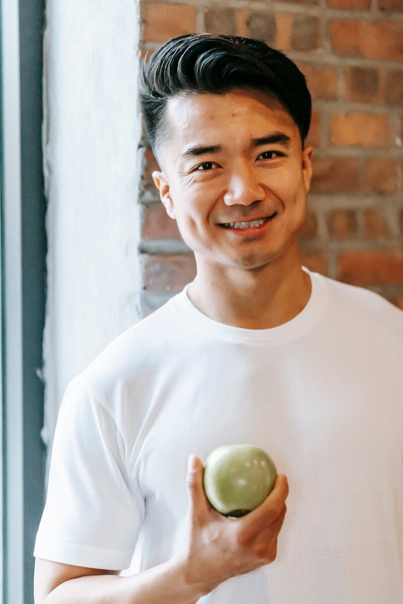 Smiling man indoors holding a green apple, promoting healthy eating and positivity.